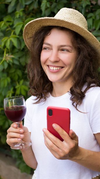Retrato de joven morena, sonriente sosteniendo copa de vino tinto, smartphone, riendo mirando a distancia.Chica feliz en sombrero de verano y camiseta blanca degustación de vino tostado en los viñedos.Vertical - Foto, imagen