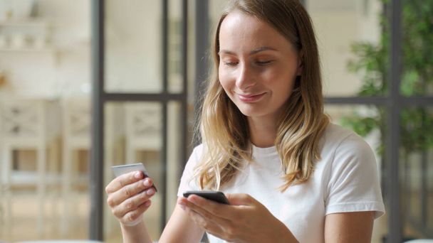 Woman holding credit card and smartphone. Young woman sitting on sofa spend money at home, make order, buying using internet, commercial services, secure on-line payment concept - Valokuva, kuva