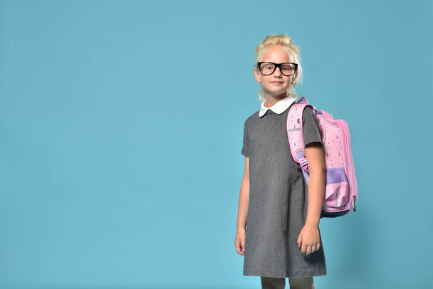 Cute preschool girl preparing for junior school and posing on a blue background with a backpack - Foto, imagen