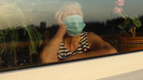 De Covid-19, gezondheid, veiligheid en pandemie concept - senior oude eenzame vrouw draagt beschermende medische masker zitten in de buurt van het raam thuis voor bescherming tegen virus - Video