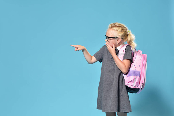 A junior schoolgirl with a backpack in surprise points her finger to the side to the free space - Foto, immagini