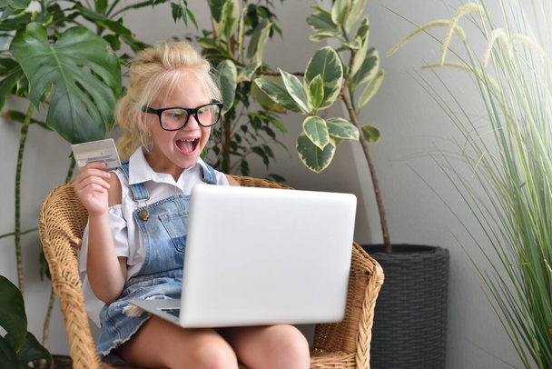 Happy little girl in glasses makes online purchases while sitting in a chair - Foto, afbeelding