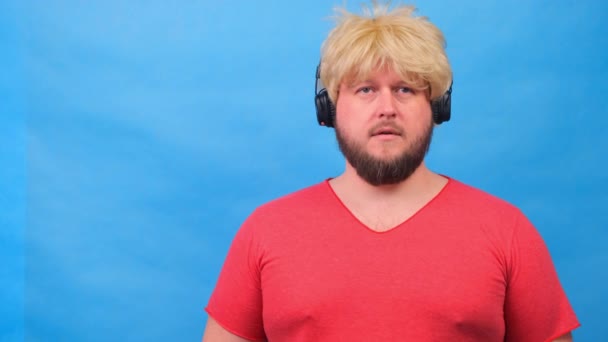 hombre gordo raro triste en una peluca y una camiseta rosa en sus manos escucha música en los auriculares y baila sobre un fondo azul - Metraje, vídeo