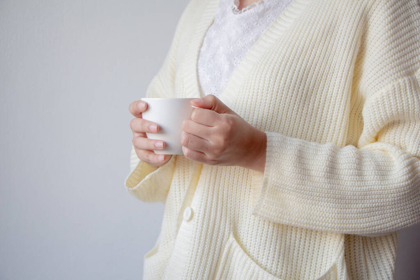 Female hand holding cup of  beverage. Beautiful girl in white sweater holding cup of tea or coffee in the morning.   - Fotó, kép