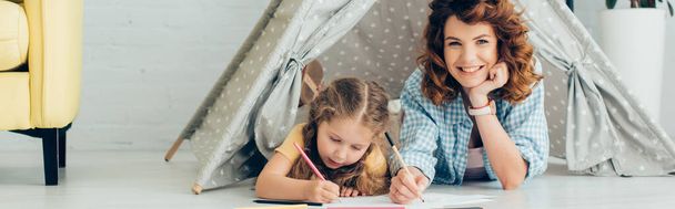 счастливая няня смотрит в камеру во время рисования вместе с ребенком в игровой палатке, панорамный снимок - Фото, изображение
