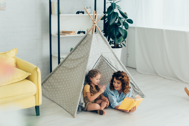 smiling babysitter reading book to adorable kid in play tent   - Foto, imagen