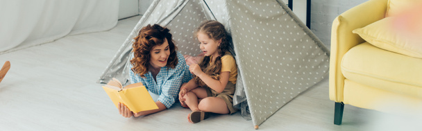 high angle view of smiling nanny reading book to adorable kid in play tent, panoramic shot - 写真・画像