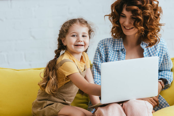 glückliches Kind schaut in die Kamera, während es Laptop zusammen mit lächelndem Babysitter benutzt - Foto, Bild