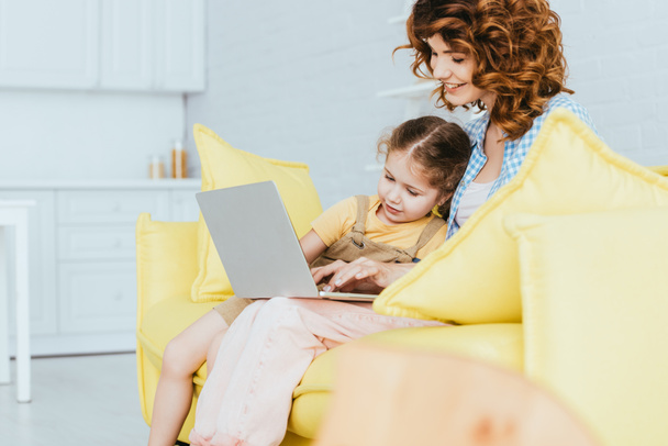 selective focus of smiling nanny and child sitting on sofa and using laptop - Valokuva, kuva