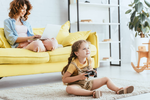 KYIV, UKRAINE - JUNE 19, 2020: adorable kid sitting on floor with joystick while young nanny using laptop on sofa - Foto, imagen