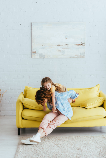 young nanny piggybacking happy kid while sitting on yellow sofa - 写真・画像