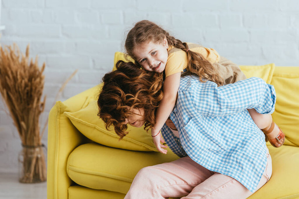 young nanny piggybacking cheerful child wihile sitting on yellow sofa at home    - Фото, зображення