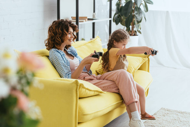 KYIV, UKRAINE - JUNE 19, 2020: selective focus of smiling babysitter and child playing video game on yellow sofa - Fotó, kép