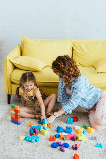 niñera joven y lindo niño jugando con bloques de construcción multicolores en el suelo cerca de sofá amarillo - Foto, imagen