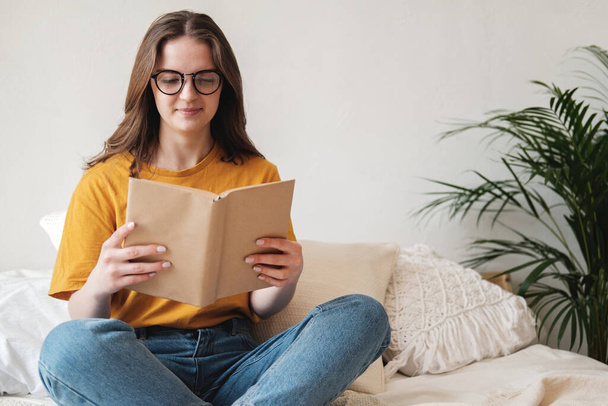 Jong mooi meisje student in bril, een oranje T-shirt en blauwe jeans zit op bed met kussens en leest papieren boek. Zelfonderwijsconcept thuis onderwijs, hobby. - Foto, afbeelding