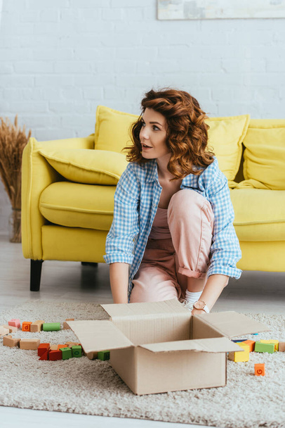 attractive young woman sitting on floor near carton box with multicolored  blocks - Foto, Bild