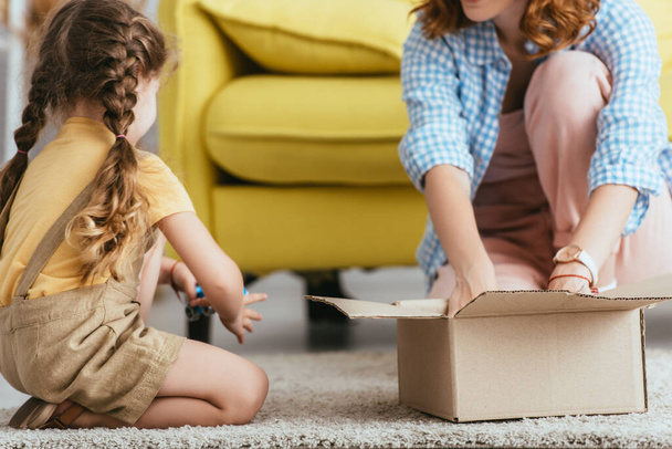 vista recortada de niñera y niño sentado en el suelo cerca de la caja de cartón - Foto, imagen