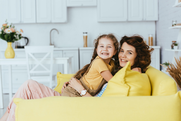 cheerful babysitter and child smiling at camera while embracing on sofa in kitchen - Photo, Image