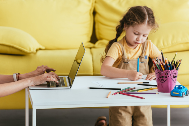 abgeschnittene Ansicht des Babysitters, der am Laptop in der Nähe von niedlichen Kinderzeichnungen mit Bleistiften arbeitet - Foto, Bild