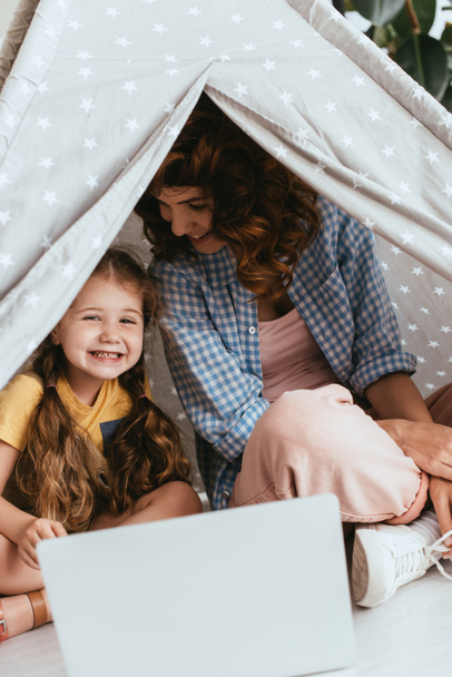 happy babysitter and adorable child sitting in toy wigwam near laptop - 写真・画像
