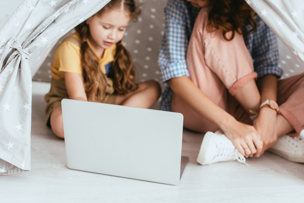 обрезанный вид няни, сидящей рядом с очаровательным ребенком с помощью ноутбука в игрушечном вигваме, избирательный фокус - Фото, изображение