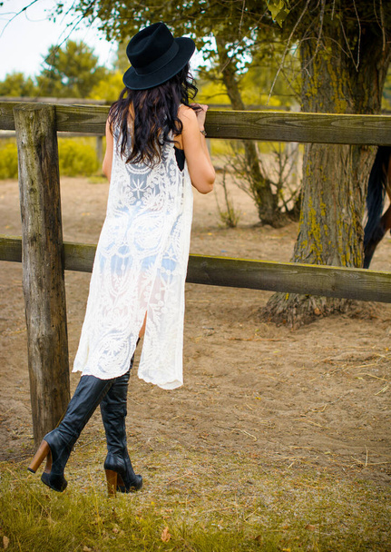Žena v černém klobouku s kudrnatými vlasy a černými šaty a s koňmi na farmě, domácí zvířata ve vesnici na ranči. Koně jsou přátelé lidí. - Fotografie, Obrázek