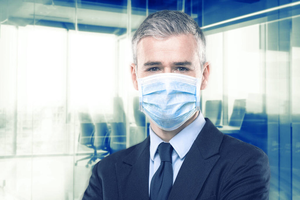 Επιχειρηματίας με κοστούμι φορώντας μάσκα προσώπου ως προστασία από το Covid-19 ή coronavirus στέκεται με διπλωμένα χέρια πάνω από ένα γκρι φόντο στούντιο - Φωτογραφία, εικόνα