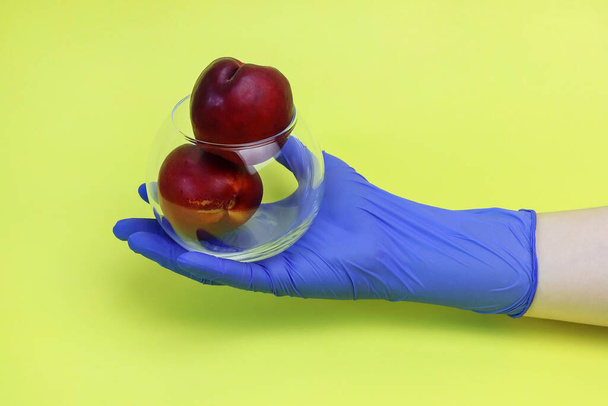 A hand in a blue glove holds a transparent dish with red nectarines on a yellow background. Copy space - concept for fresh fruit, security, quarantine, coronavirus, grocery shopping. - Фото, зображення