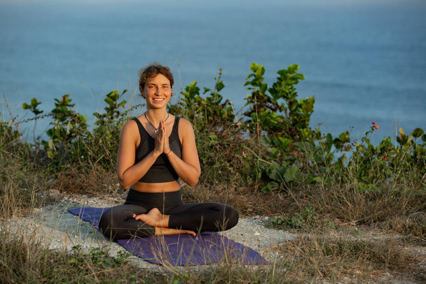 young woman sitting in lotus position and smiling at camera while practicing yoga outdoors  - Foto, Imagen