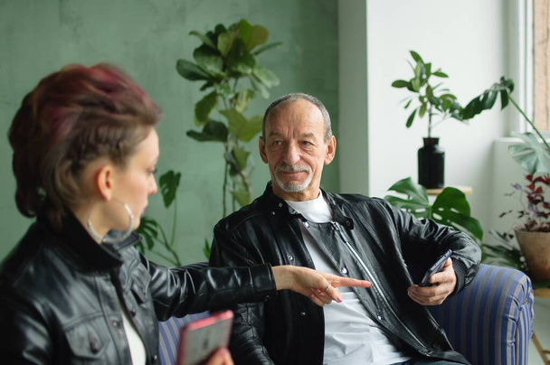 Family portrait of adult daughter and senior father in loft room with houseplants. Man and girl are wearing black leather jackets in punk style and browsing on their smartphones without communication - Foto, afbeelding