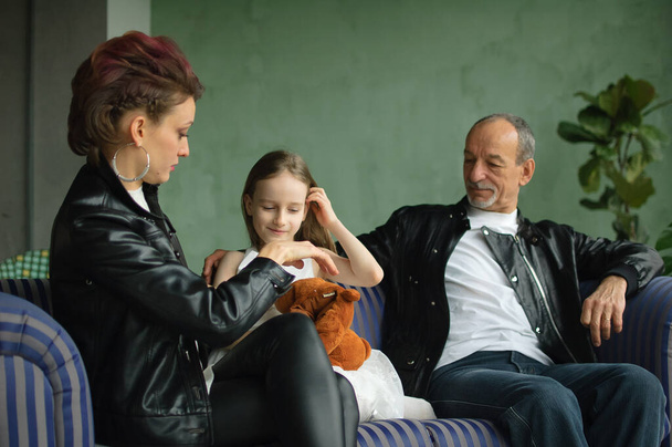 Family portrait of adult daughter, little granddaughter and senior grandfather in loft room with houseplants. Man and woman are wearing black leather jackets in punk style, generation concept - Photo, Image