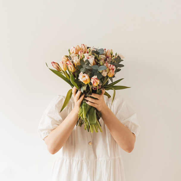 Beautiful girl in white dress hold in hands flower bouquet against white wall. Minimal beauty / fashion festive concept - Photo, Image