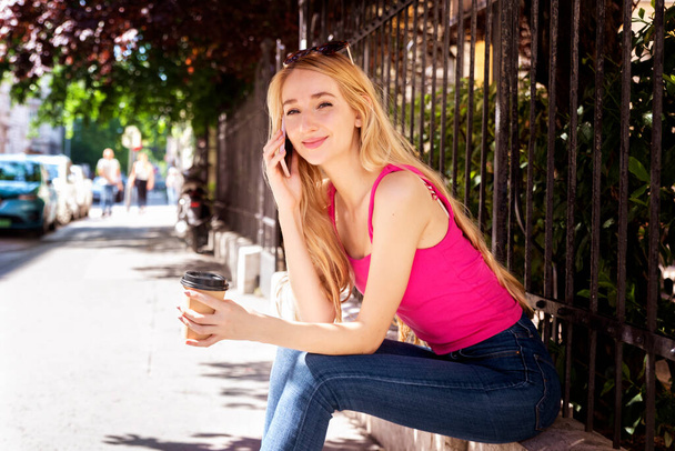 Portrait shot of smiling young woman using phone and drinking coffee while sitting on the street.  - Valokuva, kuva