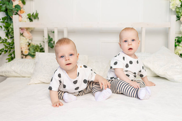 due gemelli piccoli di 8 mesi seduti sul letto con gli stessi vestiti, relazione fratello-sorella, vestiti alla moda per i figli di gemelli, concetto di matrimonio e amicizia - Foto, immagini