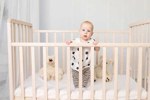 um bebê de 8 meses fica em um berço com brinquedos em pijama em um quarto de crianças brilhantes depois de dormir e olha para a câmera, um lugar para texto
 - Foto, Imagem
