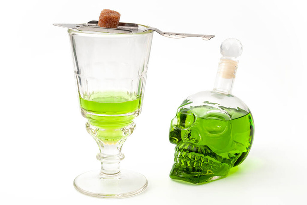 Спирт и алкоголь, зеленая фея и опасная концептуальная идея спиртного с хрустальным стаканом абсента, серебряной ложкой, сахарным кубиком и бутылкой крепкого духа в форме черепа на белом фоне - Фото, изображение