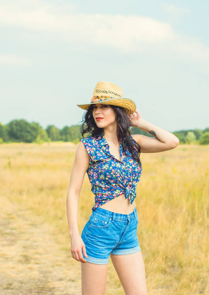Piękne gorące sexy kobieta z jasny makijaż noszenie w amerykańskim stylu country, słomy kapelusz i dżinsy szorty na złote pole tło niebo - Zdjęcie, obraz