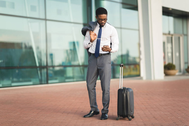 Чорний підприємець, який користується мобільним телефоном, стоїть з багажем в аеропорту. - Фото, зображення