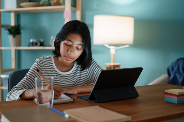 Homeschooling chica asiática haciendo la tarea y estudiar en línea con la tableta en un escritorio por la noche
. - Foto, imagen