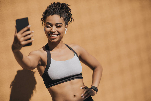 African american young woman in sports bra with wireless headphones and fitness tracker making selfie - Foto, immagini