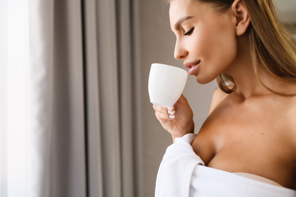 お茶を飲みながら窓の近くに柔らかい肌を持つ美しい若い女性は、シャワー後のコーヒー、背景にグレーのカーテンで白いタオルを身に着けています。顔の官能的なブロンド女性で幸せな朝. - 写真・画像