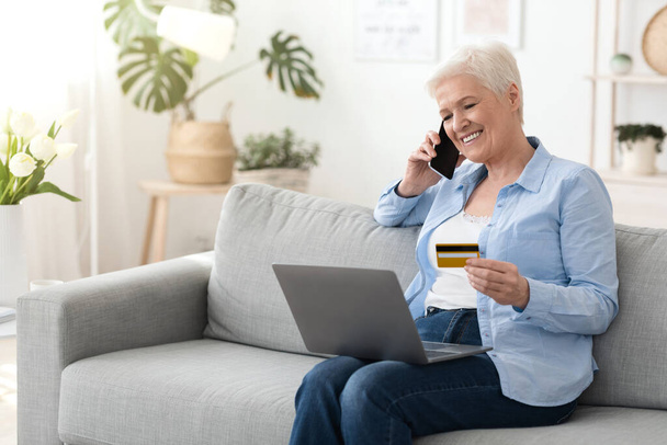 Femme âgée avec ordinateur portable et carte de crédit parlant sur téléphone portable à la maison - Photo, image