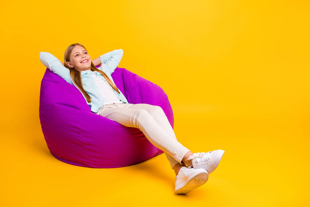 Full length photo of beautiful little blond lady good mood hold hands behind head sitting lying cozy soft violet bean bag wear casual shirt trousers footwear isolated yellow color background - Photo, Image