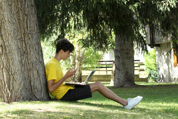 adolescente masculino utilizar un ordenador portátil acostado en la hierba apoyada contra un árbol con espacio de copia para su texto
 - Foto, Imagen