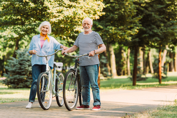 Couple âgé souriant à la caméra tout en marchant avec des vélos sur le trottoir dans le parc  - Photo, image