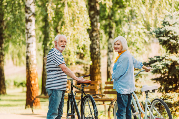 Senior couple smiling at camera while standing near bicycles in park - Фото, изображение
