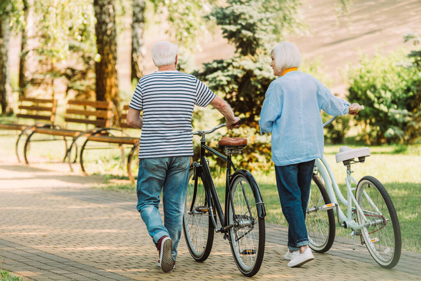 Widok z tyłu para seniorów spacerujących w pobliżu rowerów w parku  - Zdjęcie, obraz