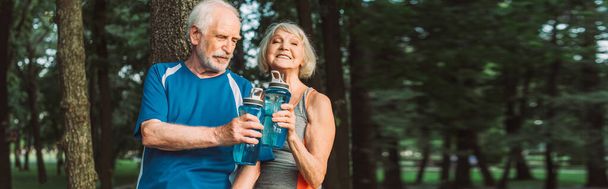 Панорамный снимок улыбающейся пожилой женщины, держащей спортивную бутылку рядом с мужем в парке  - Фото, изображение