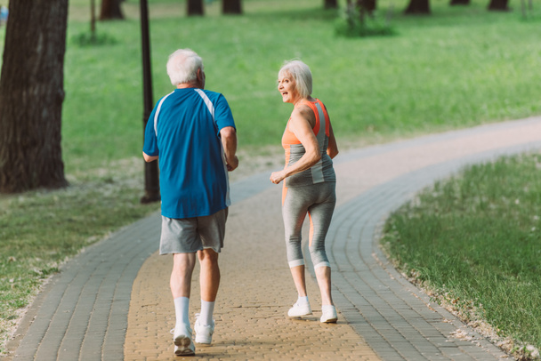 Back view of smiling senior woman looking at husband while jogging in park  - Foto, Imagem