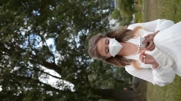 Elegant stylish woman in a white dress and a protective mask on her face sits on the lawn in the park and looks into her smartphone - Video